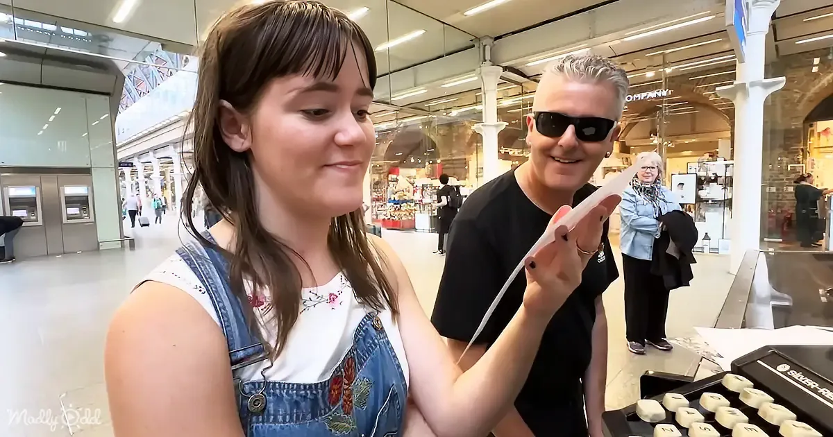 A woman in denim overalls holds a white object, smiling, with a man in sunglasses standing beside her in a busy indoor...