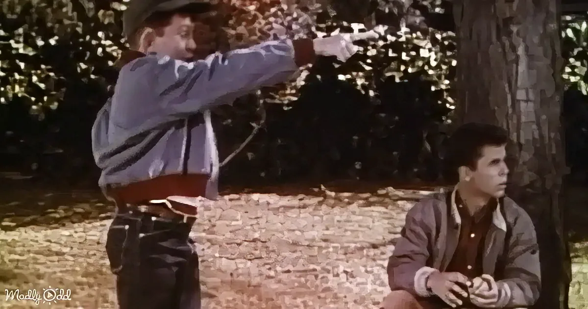 A man in a cowboy hat points a gun while another man sits nearby in a forest setting.