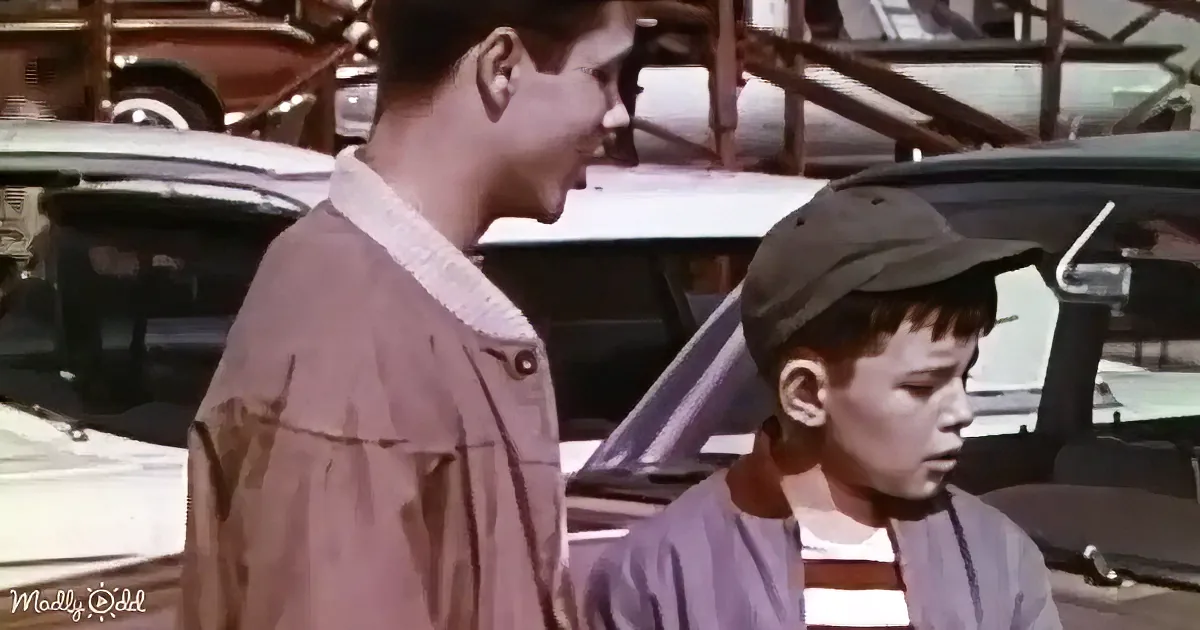 A young boy in a baseball cap stands beside an older boy in a brown shirt, both in a parking lot with vintage cars.