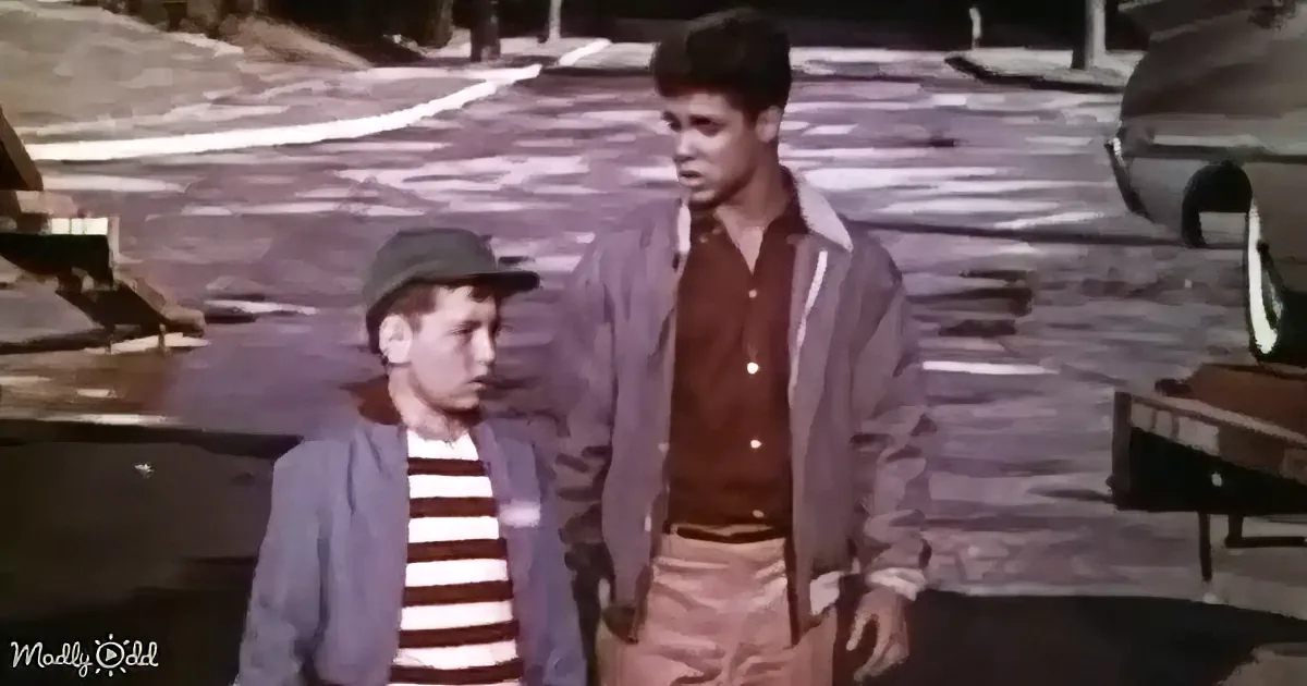 A young boy in a striped shirt and cap stands next to an older boy in a jacket and brown shirt, both on a street with...