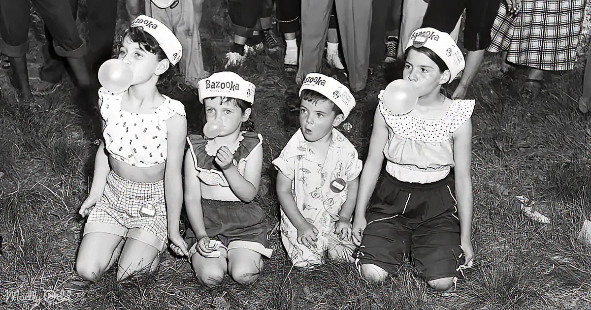 Four children in Bazooka bubble gum hats sit on grass, two blow bubbles, one looks surprised.