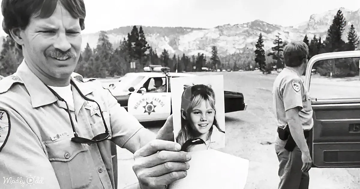A man in a sheriff's uniform holds a photo of a smiling girl, with a police car and another officer in the background.