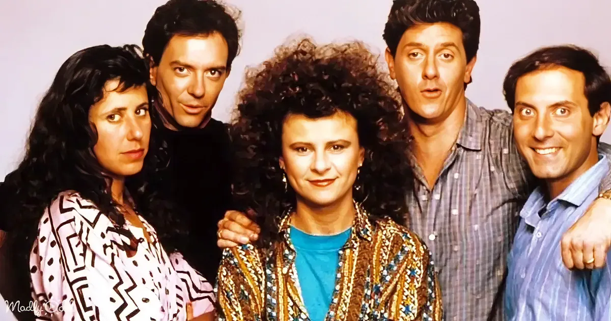 A group of five people, dressed in 80s fashion, pose together with one person in the center.