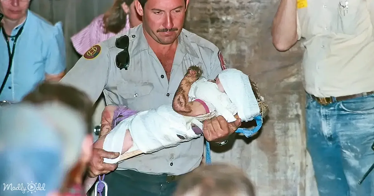 A man in a uniform holds a baby wrapped in white cloth, surrounded by onlookers.