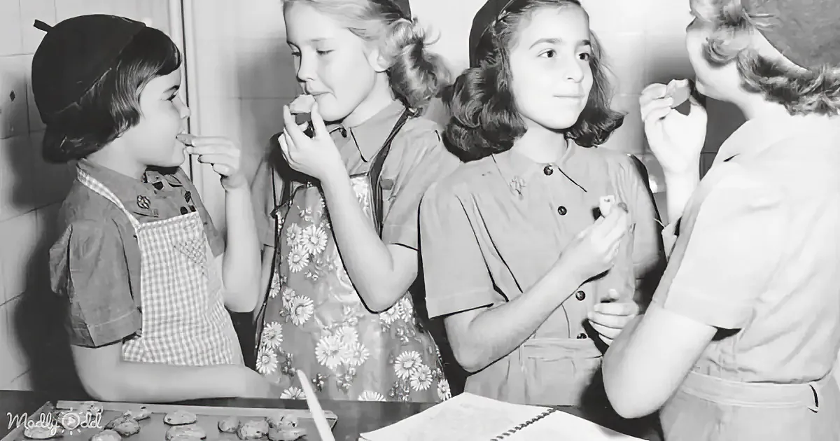 Four girls in aprons are eating cookies, with one feeding another, in a kitchen setting.