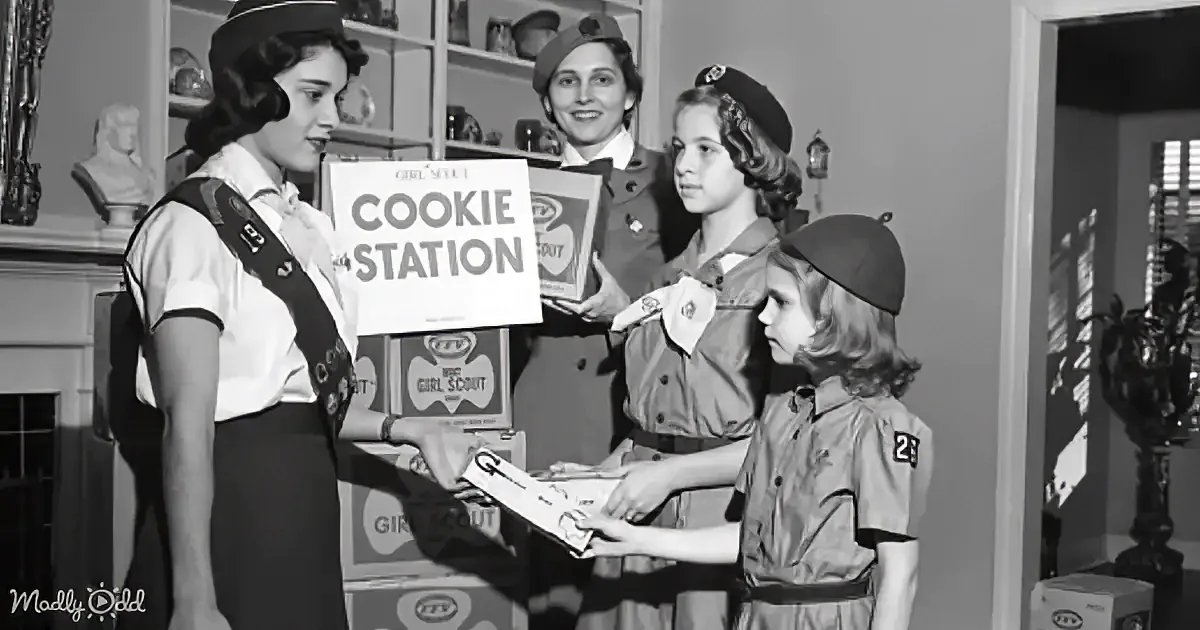 A Girl Scout holds a "Cookie Station" sign while interacting with other scouts in a vintage setting.