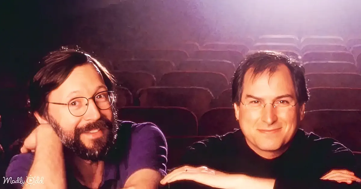 Two men are sitting in a dimly lit theater, one with glasses and a beard, the other with short hair, both smiling.