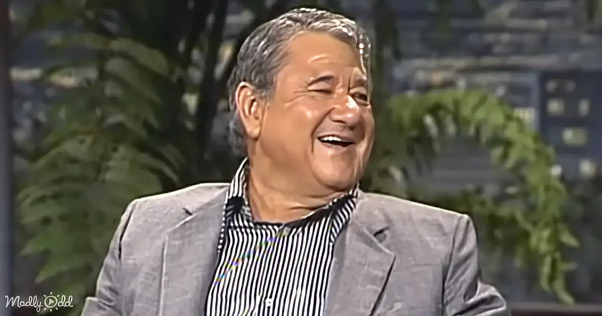 A man in a striped shirt and gray blazer is smiling, with greenery and a cityscape in the background.