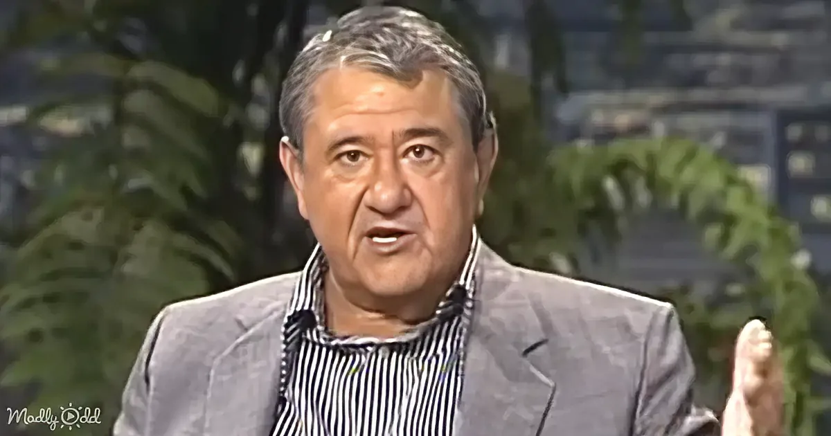 A man in a striped shirt and gray blazer is speaking, with a blurred background of greenery.