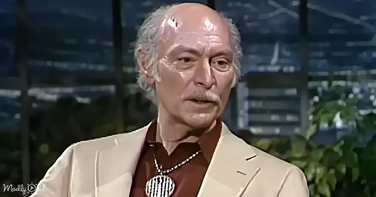 A man with a bald head, mustache, and glasses, wearing a beige suit and a brown shirt with a silver necklace, is speaking.