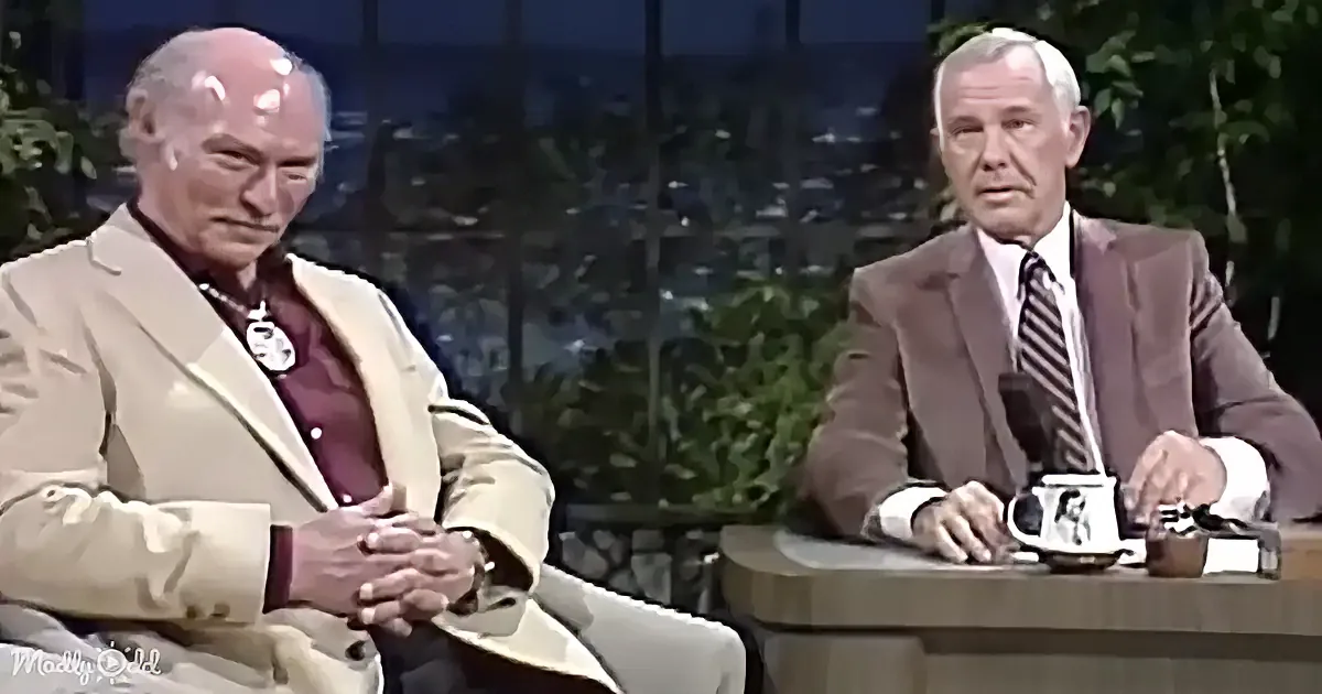 Two men in suits are seated, one in a beige suit with a necklace, the other in a brown suit with a striped tie.