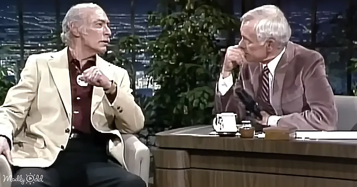 Two men in suits are seated, one gesturing while the other listens, in a talk show setting with a cityscape backdrop.
