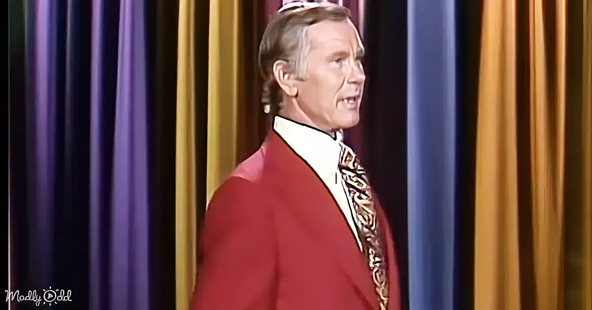 A man in a red suit and gold tie stands against a colorful curtain backdrop.