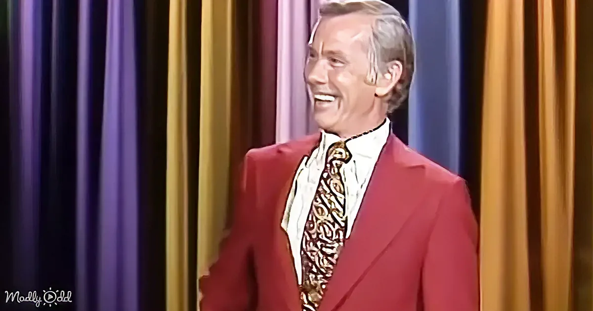 A man in a red suit and patterned tie smiles on a stage with colorful curtains.