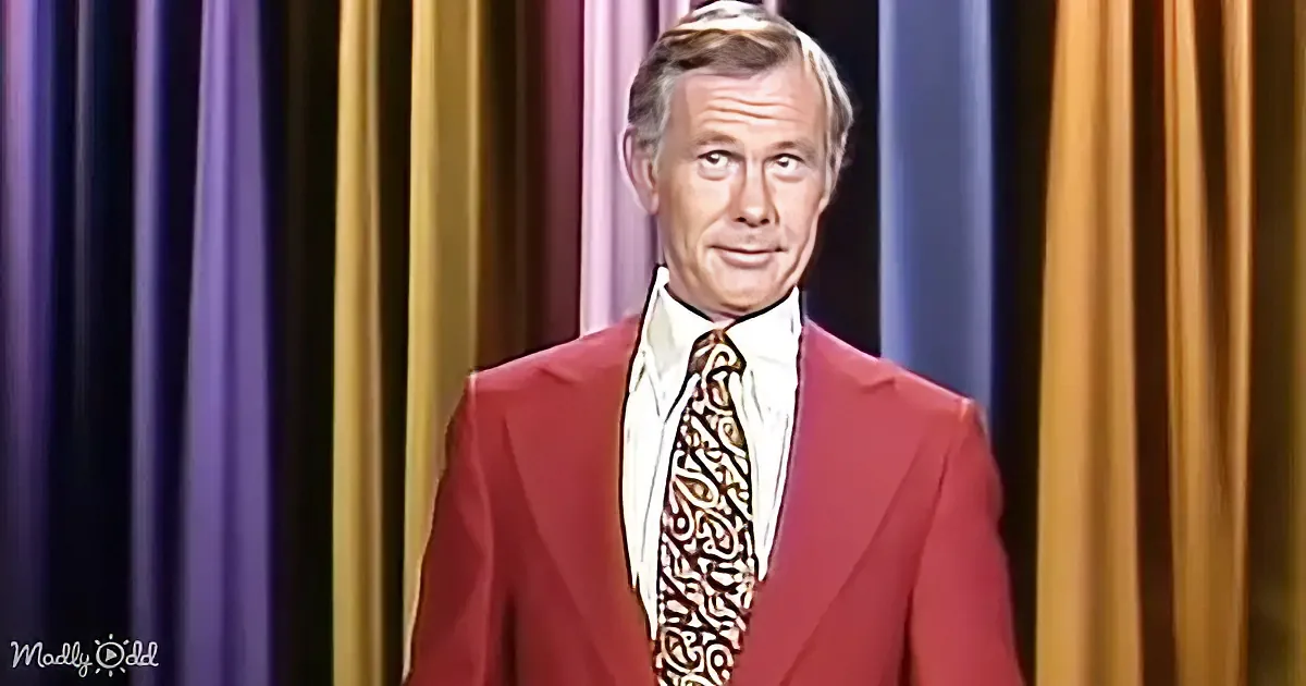 A man in a red suit and patterned tie stands in front of a colorful curtain backdrop.