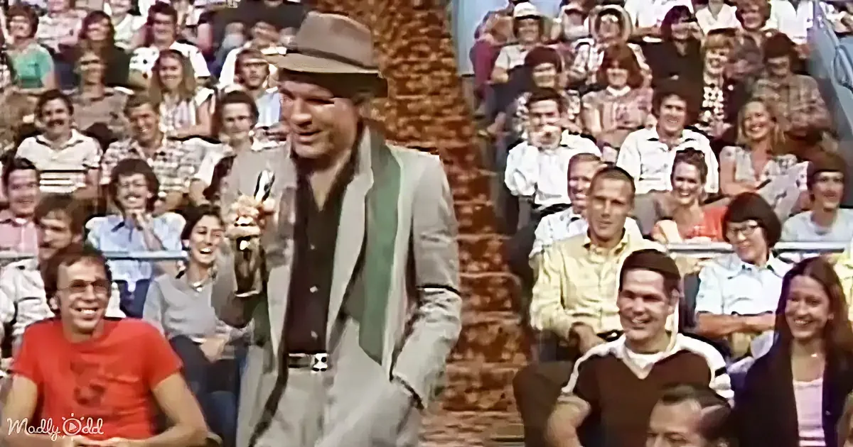 A man in a hat and suit holds a trophy, smiling, with a crowd of people in the background.