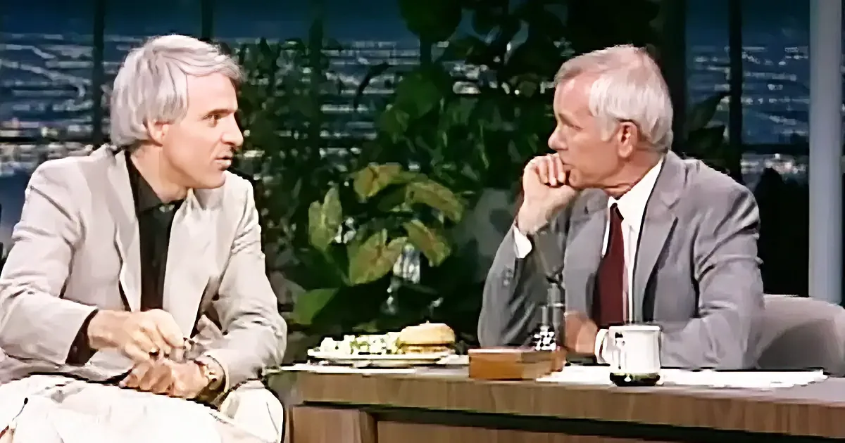 Two men in suits are seated at a table, engaged in conversation, with a cityscape and plants in the background.