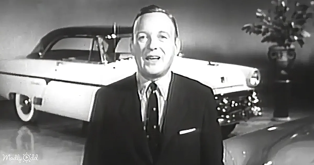 A man in a suit stands in front of a vintage car, smiling, with a vase of flowers nearby.
