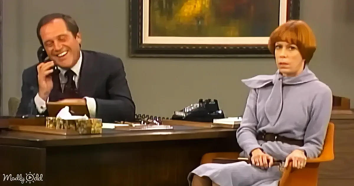 A man in a suit laughs on the phone while a woman in a blue dress sits nearby, both in an office setting.