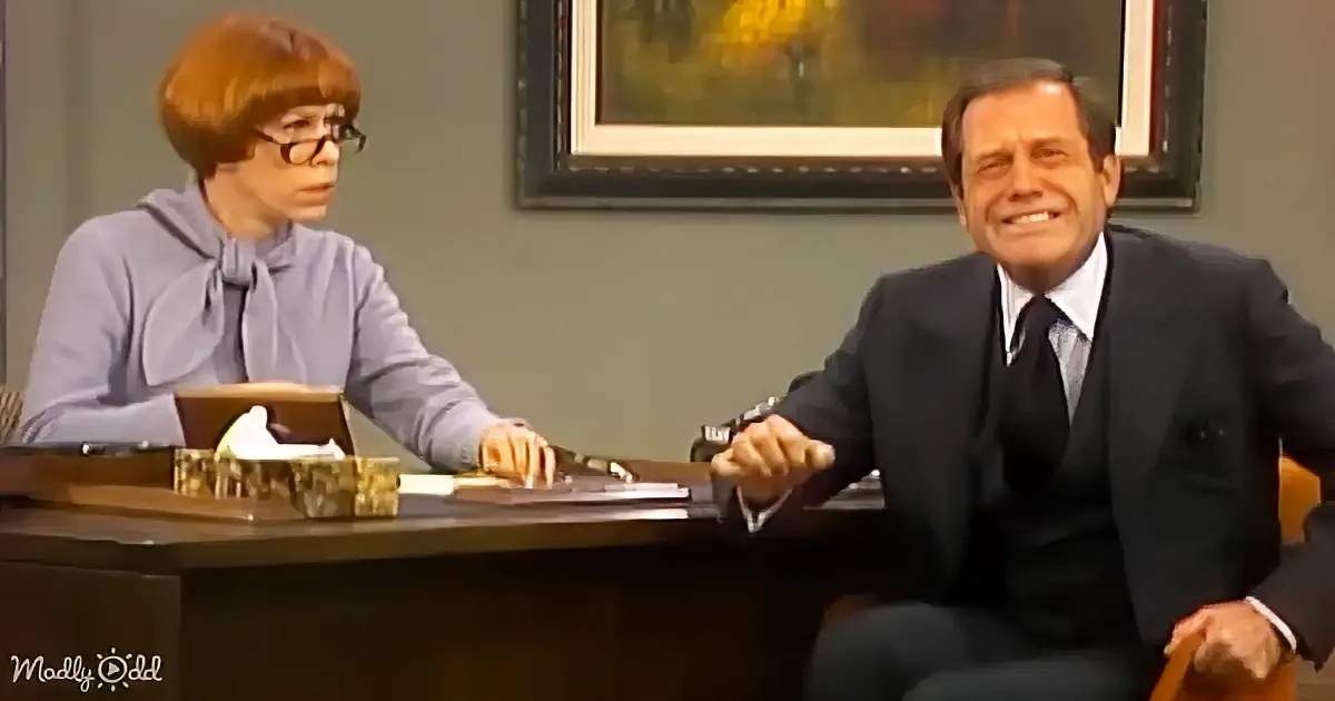 A man in a suit sits at a desk, smiling, while a woman in a purple top and glasses looks on.