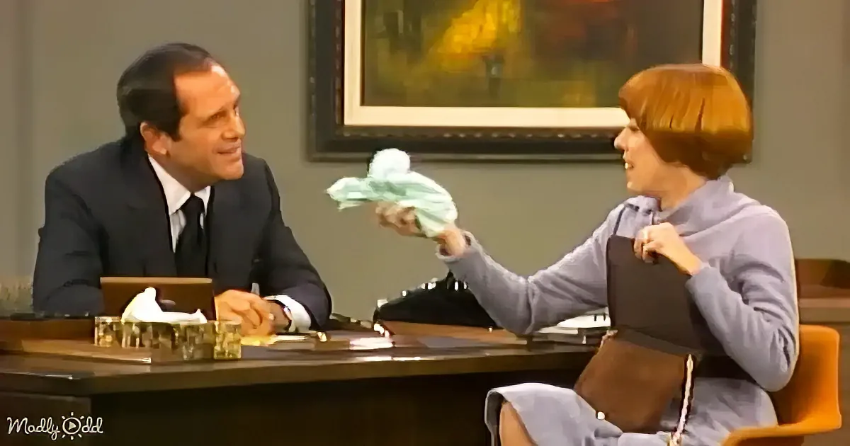 A woman in a purple outfit holds a green cloth while seated at a desk with a man in a suit, both facing each other.