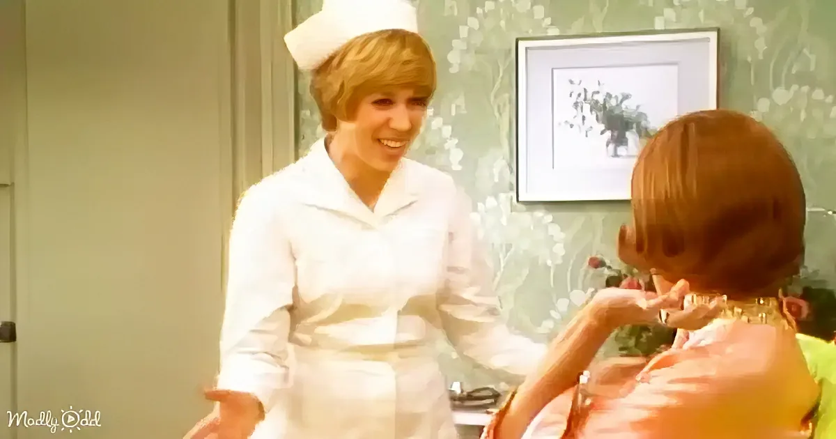 A nurse in white uniform smiles at a woman in a pink dress, both in a room with floral wallpaper.