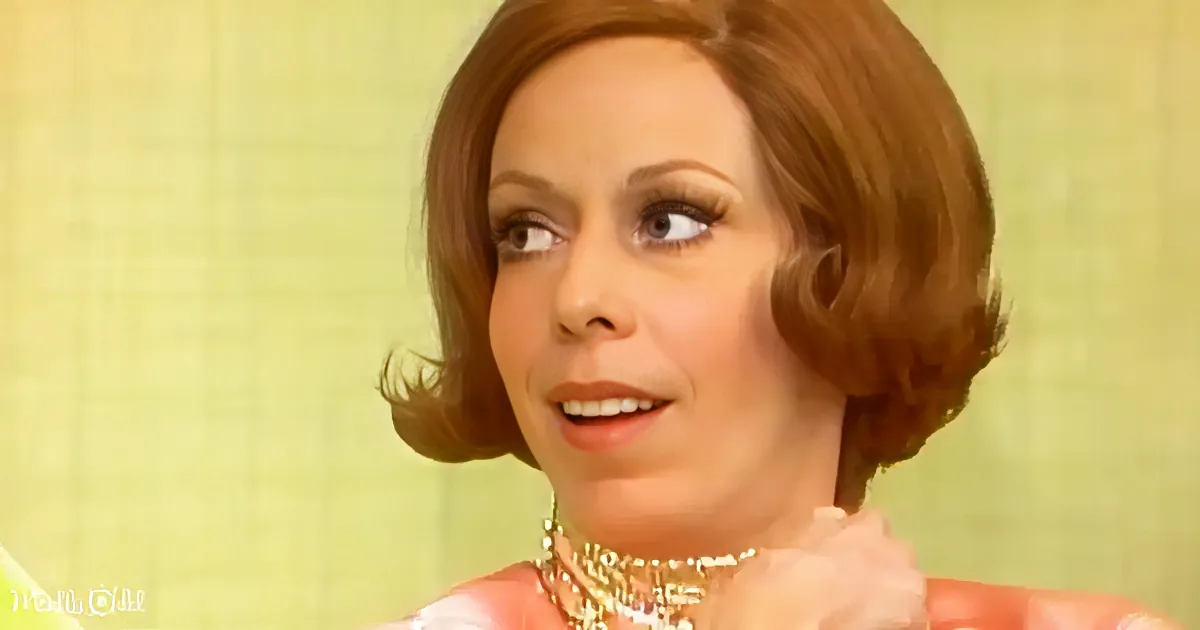 A woman with short, styled brown hair, wearing a pink outfit and a beaded necklace, looks to the side with a slight smile.