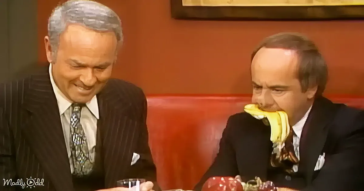 A man in a suit is eating a banana while another man in a suit looks on, both seated at a table.