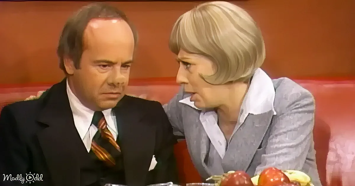 A man in a suit and tie sits next to a woman with blonde hair, both appearing to be in a formal setting.