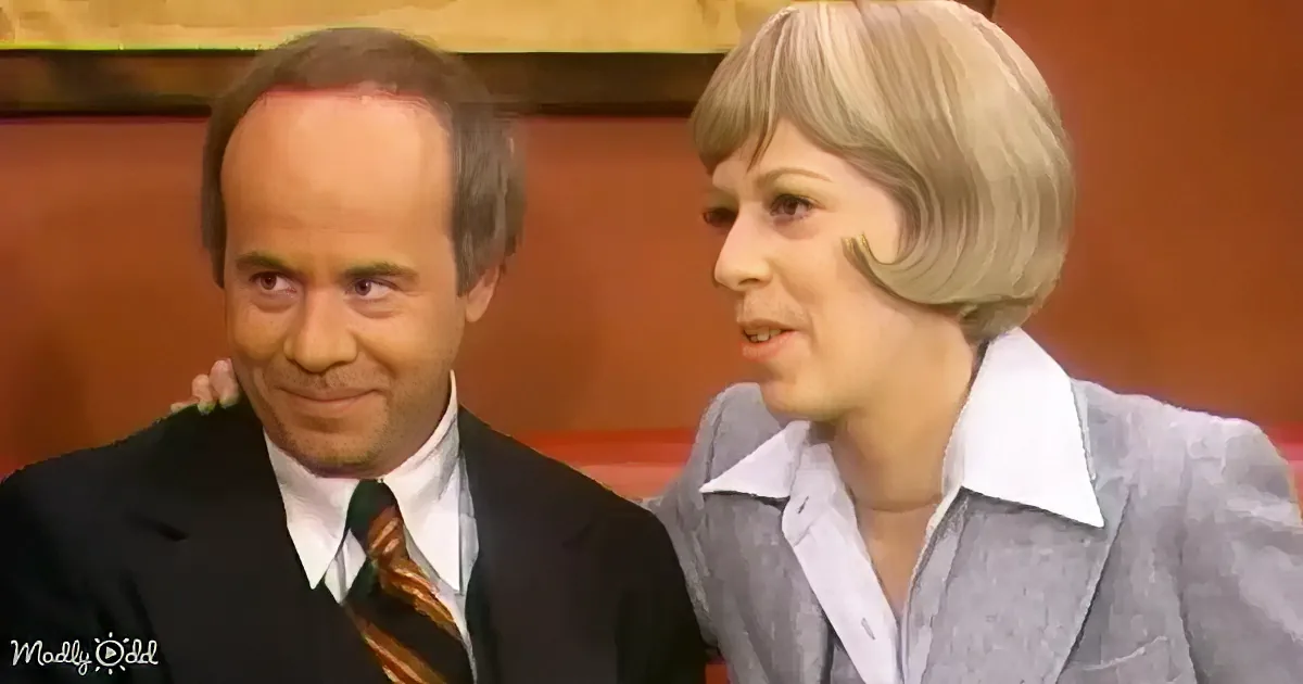 A man in a suit and tie sits next to a woman in a gray blazer, both appearing to be in a studio setting.