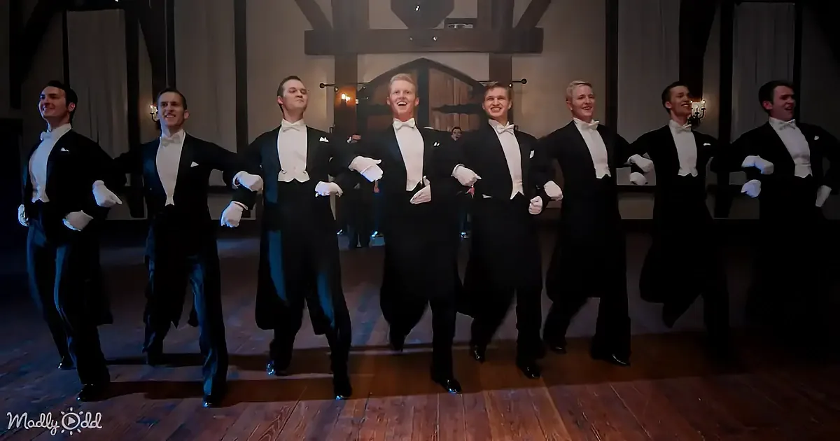 A group of men in tuxedos and white gloves are dancing in a dimly lit room with wooden floors and exposed beams.