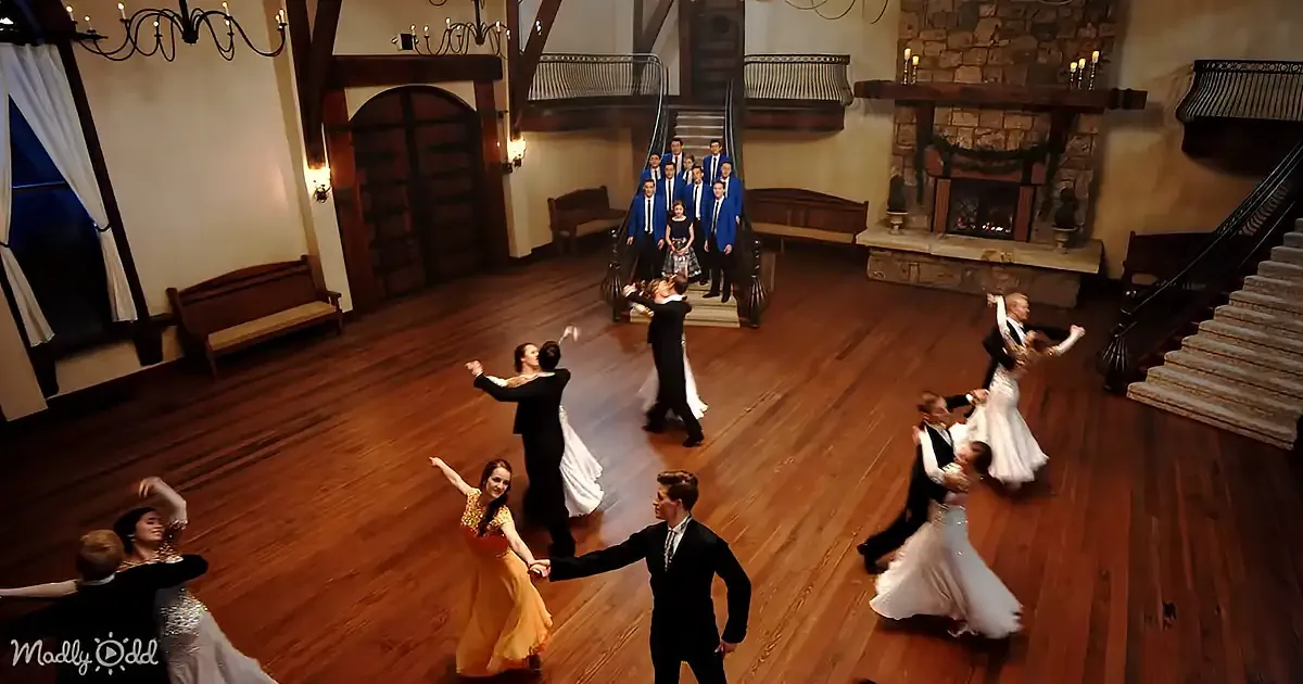 A group of people in formal attire dances gracefully in a grand, warmly lit room with wooden floors and a stone fireplace.