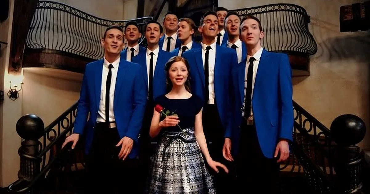 A woman in a black and white dress holds a rose, standing on a staircase with a group of men in blue suits.