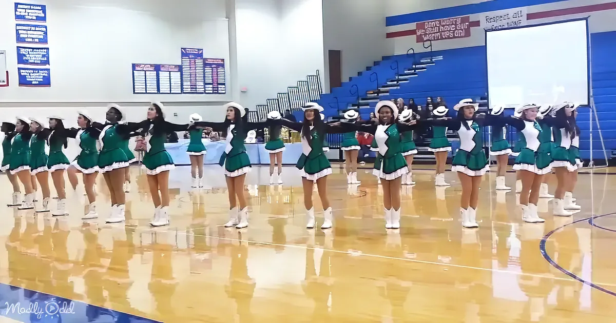 Cheerleaders in green uniforms perform a synchronized routine on a polished gym floor.
