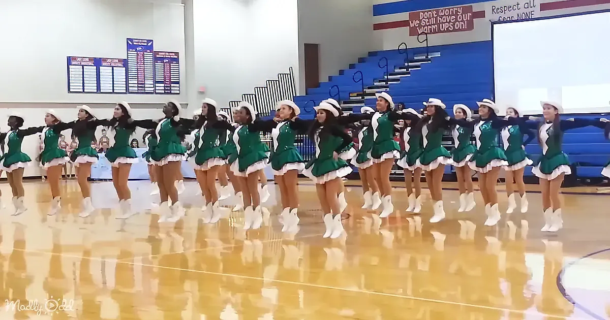 A synchronized dance team in green uniforms performs on a polished gym floor.