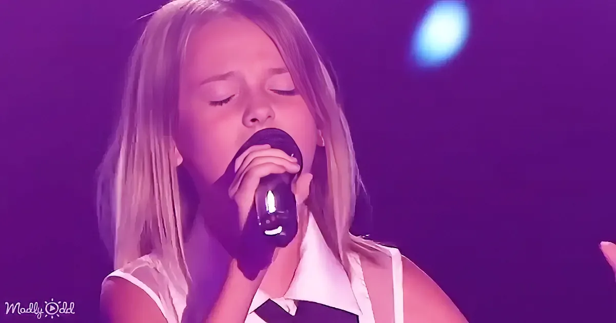 A person singing into a microphone with eyes closed, wearing a white top, under purple stage lighting.
