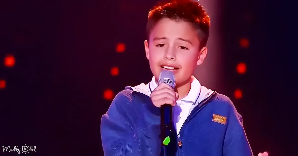 A young boy in a blue jacket sings into a microphone on stage with red lights in the background.