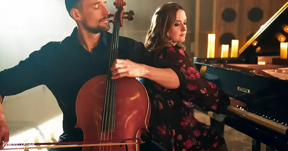A man plays cello while a woman in a floral dress sits beside him, both in a warmly lit room.