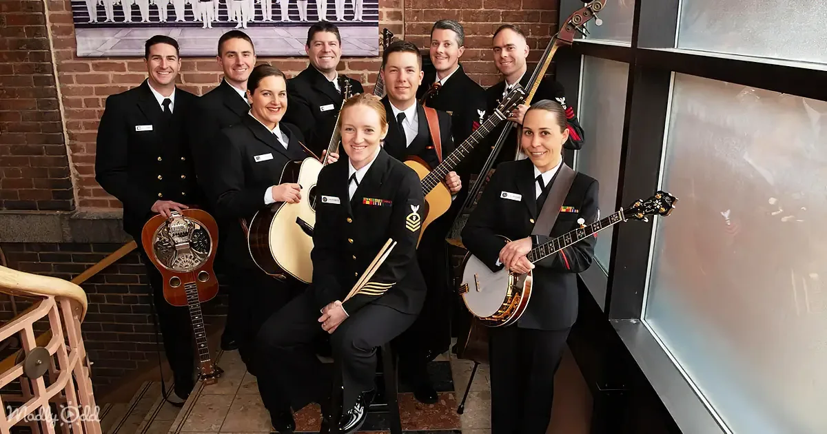 A group of musicians in uniform pose with various string instruments in a brick-walled setting.