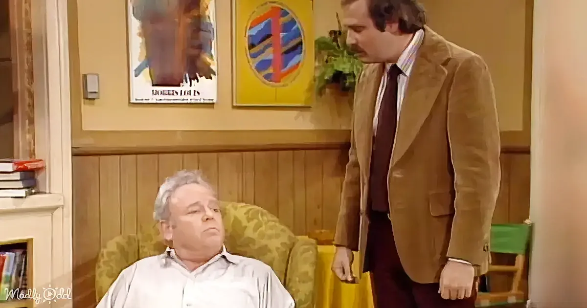 A man in a suit stands beside another man in a chair, both in a room with wood paneling and framed pictures.