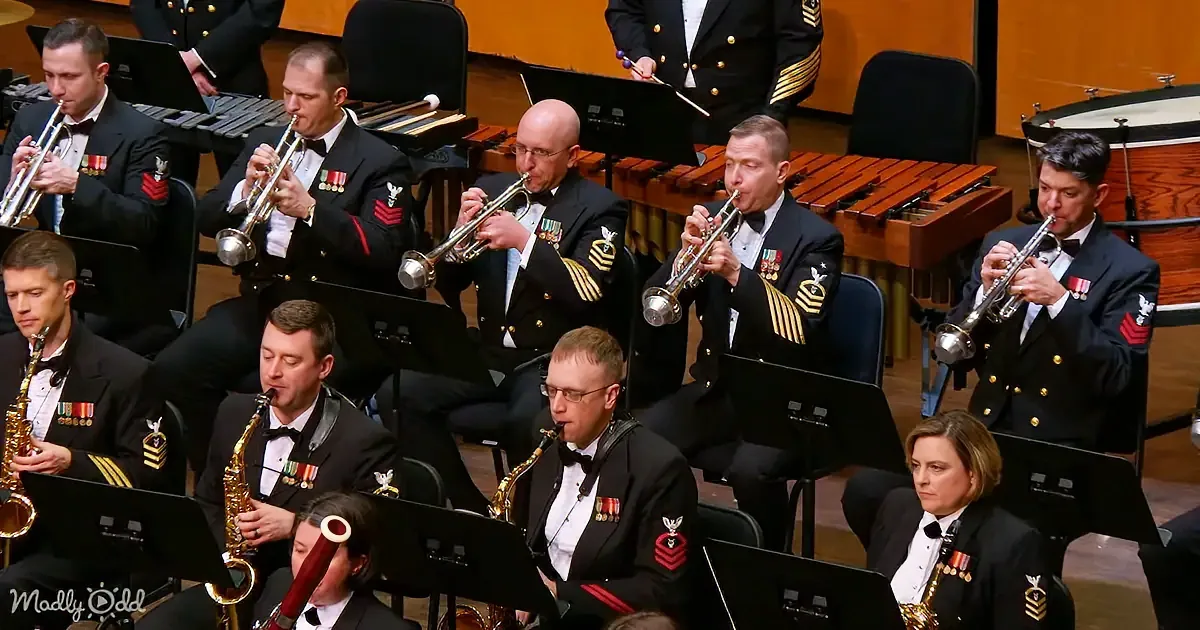 A military band in formal uniforms plays brass and woodwind instruments in a concert setting.