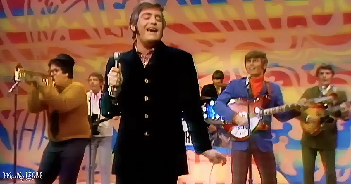 A man in a black coat sings passionately on stage with a colorful band behind him.