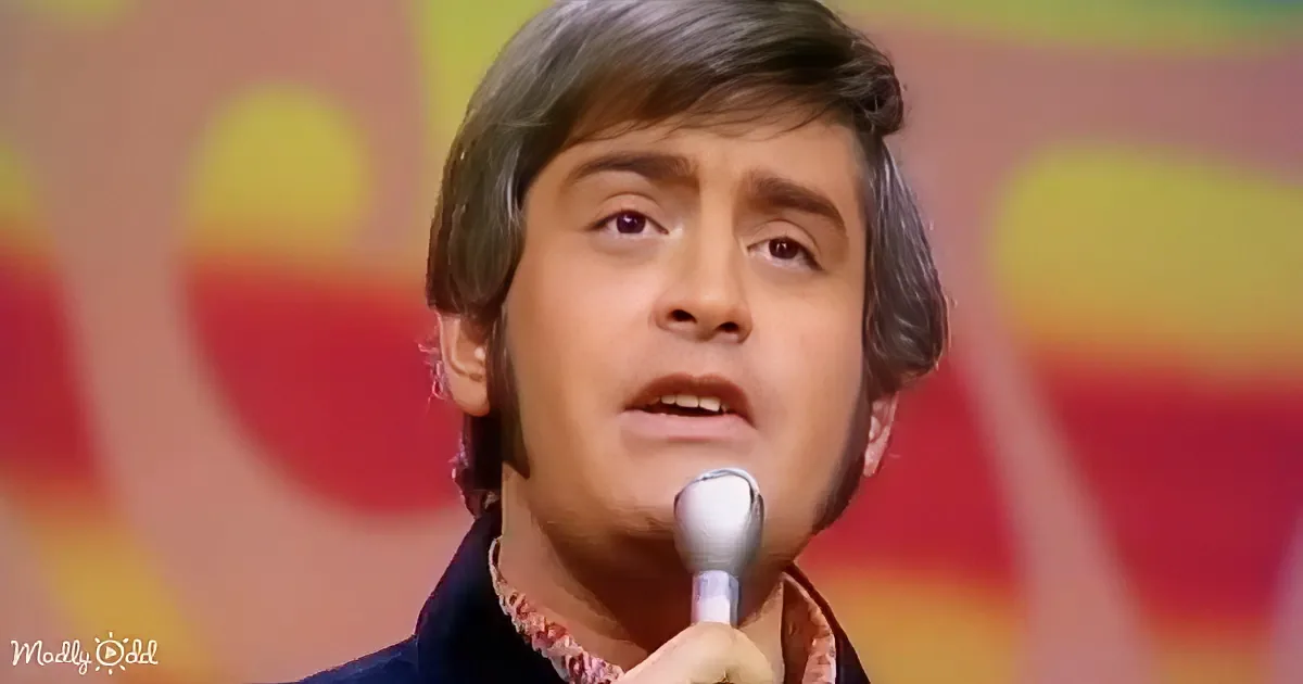 A man with dark hair and a patterned shirt holds a microphone, singing against a colorful background.