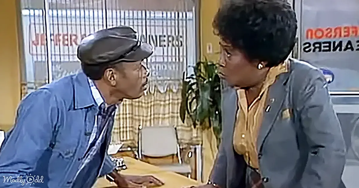 A man in a cap leans toward a woman in a gray jacket, both seated at a table in a room with a window and a "Jeepers" sign.