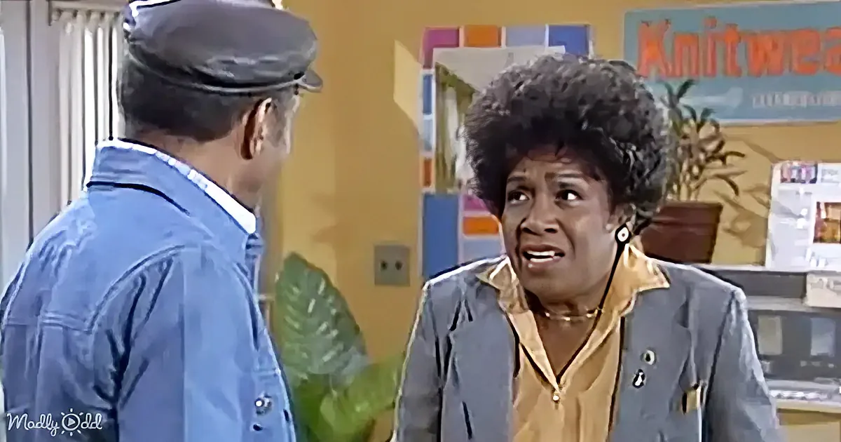 A woman with curly hair and a blue jacket is speaking to a man in a blue shirt and cap in a room with a "Knitwear" sign.