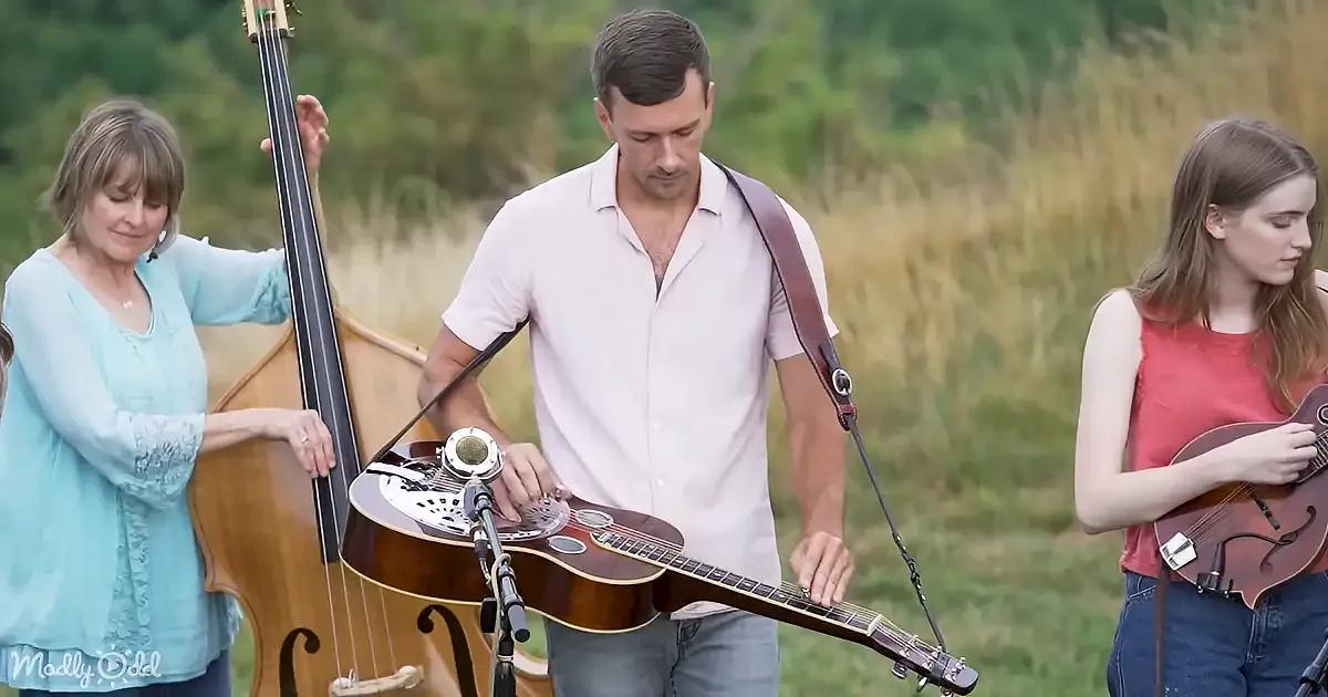 A man plays a resonator guitar in a field, accompanied by a woman on a double bass and another woman on a mandolin.