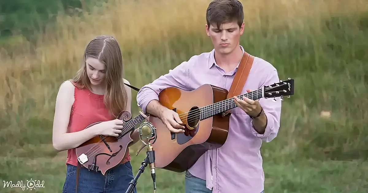 A man and woman play acoustic guitar and mandolin in a grassy field.