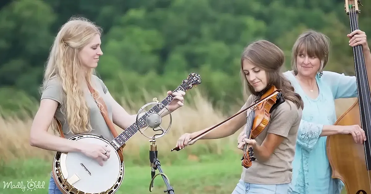 A woman plays a banjo while another holds a violin, and a third person stands with a bass, all in a lush green field.