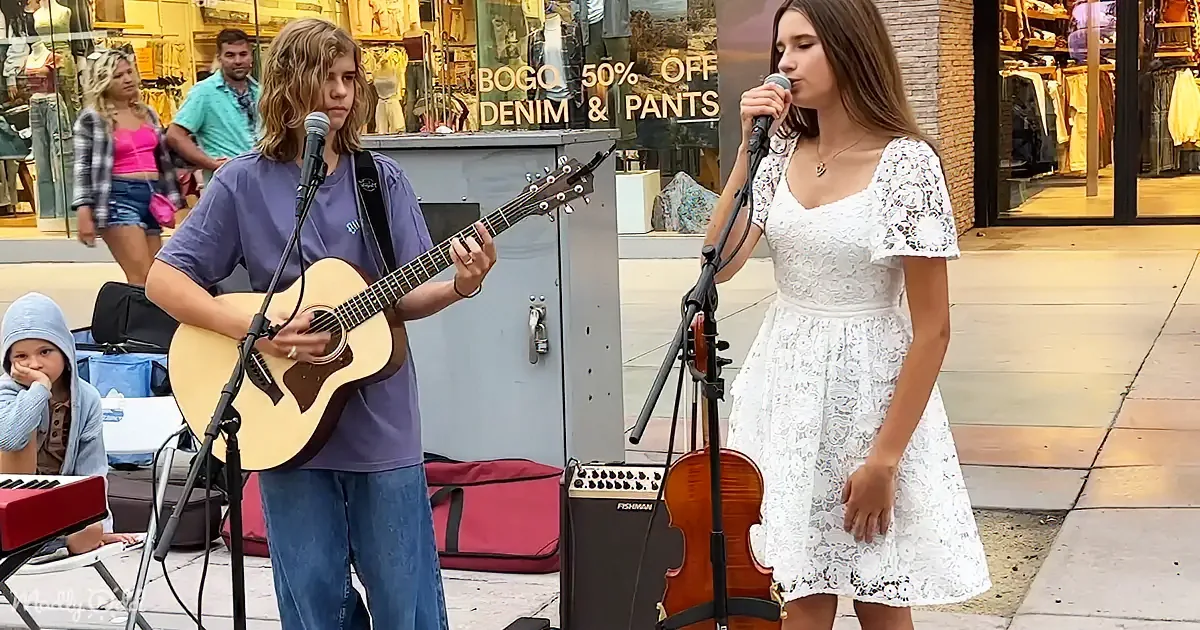 A young man plays guitar while a young woman sings into a microphone on a sidewalk.