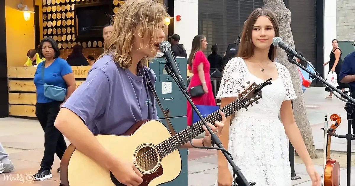 A young man plays guitar and sings, accompanied by a young woman in a white dress, both on a sidewalk with passersby.
